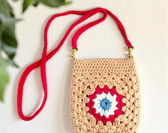 May include: A beige crochet crossbody bag with a red strap and gold-colored clasps. The bag features a central square design with red, white, and blue detailing, creating a floral pattern. The bag is handmade.