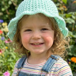 Puede incluir: Un sombrero de sol de ganchillo verde claro con un borde festoneado. El sombrero lo lleva un niño pequeño con un peto vaquero y una camiseta de rayas. El niño sonríe frente a un jardín colorido.