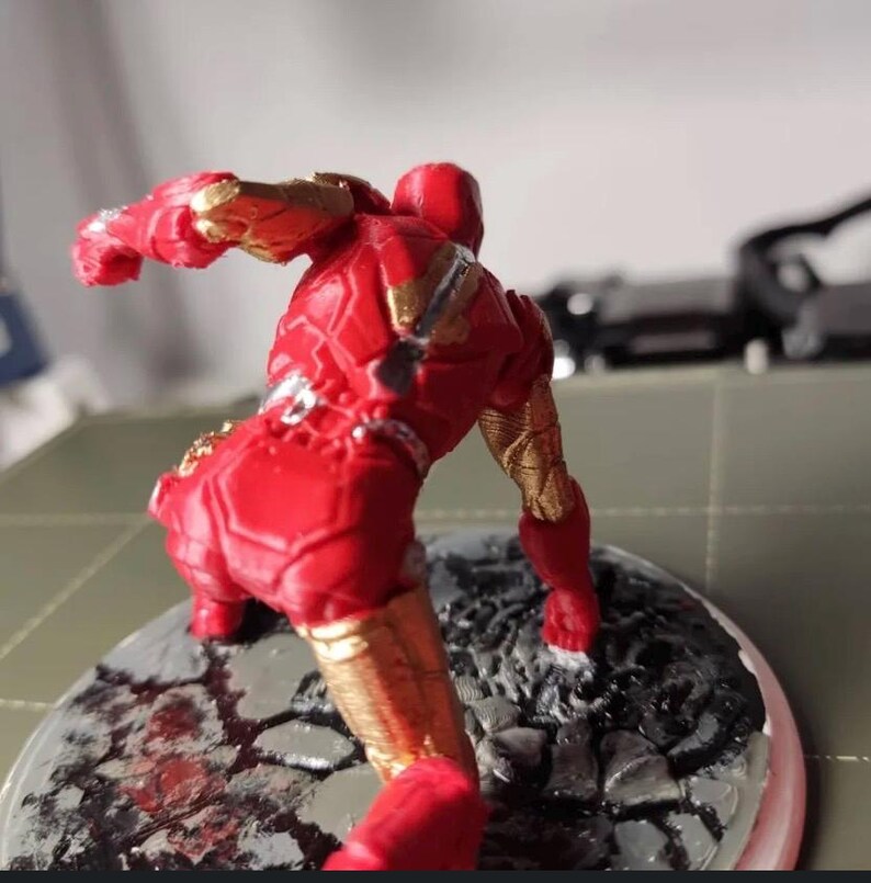 May include: A red and gold Iron Man action figure, posed in a dynamic stance. The figure is detailed with metallic gold accents and stands on a circular base with a textured, rocky surface. The figure is a collectible item.