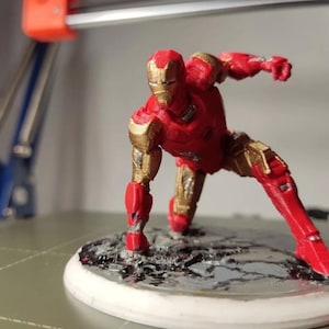 May include: A red and gold Iron Man figurine in a dynamic pose, with one arm extended. The figurine is on a circular base with a textured surface. The figure is approximately 5 inches tall.