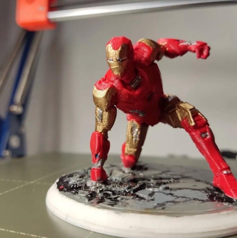 May include: A red and gold Iron Man figurine in a dynamic pose, with one arm extended. The figurine is on a circular base with a textured surface. The figure is approximately 5 inches tall.