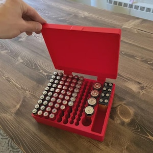 Puede incluir: Un organizador de baterías rojo, abierto para mostrar varios tamaños de baterías. El organizador está lleno de pilas AA, AAA, C, D y de 9 voltios. La caja es de plástico y tiene una tapa con bisagras.