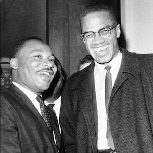 May include: Black and white photograph of Martin Luther King Jr. and Malcolm X shaking hands. Both men are wearing suits and ties. King is smiling and has a friendly expression. X is also smiling and appears to be laughing.