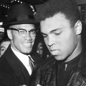 May include: A black and white photograph of two men, one wearing a hat and the other wearing a leather jacket. The man in the hat is smiling and the man in the jacket is looking down.