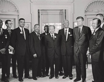 Op de afbeelding: Een zwart-witfoto van een groep mannen in pak, waaronder Martin Luther King Jr. en president John F. Kennedy, die samen in een kamer staan.