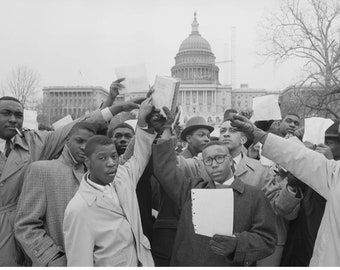 May include: A group of young Black men stand in front of the US Capitol building, holding up papers and a book. They are protesting for civil rights.