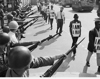 Op de afbeelding: Een zwart-witfoto toont een rij soldaten met helmen die geweren richten op een groep mensen die meelopen in een protest. De individuen dragen witte borden met de vetgedrukte zwarte tekst I AM A MAN. Militaire voertuigen zijn zichtbaar.