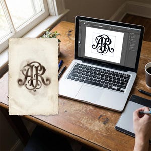 May include: A vintage-style monogram design featuring the letters "AR" in an ornate, swirling script. The design is displayed on a piece of aged paper and on a laptop screen, suggesting digital design work. The scene includes a wooden desk, a laptop, and design tools.