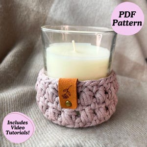 May include: A crocheted candle cozy in a dusty rose color with a brown leather strap and a small flower design. The cozy is around a clear glass candle jar with a white candle inside. The text "PDF Pattern" is in a pink circle in the upper right corner of the image. The text "Includes Video Tutorials!" is in a pink circle in the lower left corner of the image.