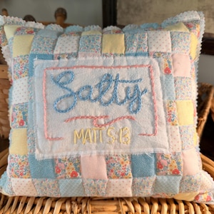 May include: Square patchwork pillow with the word "Salty" embroidered in blue script. The pillow features a mix of pastel floral and solid fabric squares, with a scalloped edge. The name "Mattie" is embroidered below "Salty".
