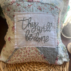 May include: A square quilted throw pillow featuring a patchwork design of blue and white floral and gingham fabrics. A central white fabric square is appliqued onto the front, featuring the embroidered dark grey text "thy will be done".