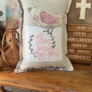 May include: A decorative pillow with a beige and white striped fabric cover. The pillow features an embroidered bird with the words "then sing my soul" in pink thread. The pillow is sitting on a wooden surface with a wooden sign with the words "Let God's Promises Shine On Your Problems" in white letters.