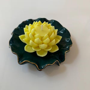 Może przedstawiać: Ż&oacute;łta świeca w kształcie kwiatu lotosu na ciemnozielonej ceramicznej podstawce z pozłacanym brzegiem.