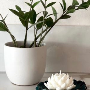 Może przedstawiać: Biała ceramiczna świeca w kształcie kwiatu lotosu stoi na ciemnozielonej ceramicznej podstawce z falistym brzegiem. W tle znajduje się doniczkowa roślina.