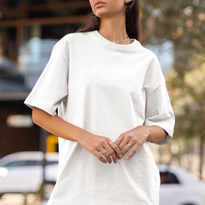 Könnte beinhalten: Ein weißes Oversize-T-Shirt mit kurzen Ärmeln. Das Model trägt das T-Shirt mit einer blauen Jeans-Shorts und einer schwarzen Sonnenbrille.