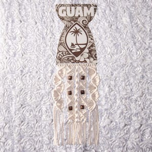 May include: A macrame wall hanging featuring a wooden sign with the word "GUAM" at the top. The sign has a brown and white design with a palm tree and boat illustration. The macrame is cream-colored with wooden beads.