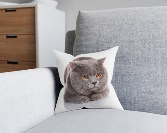 May include: A square white throw pillow featuring a high-resolution photographic print of a grey British Shorthair cat with bright orange eyes. The cat is shown in a resting position with its paws tucked underneath its body on a plain background.