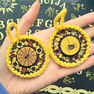 Puede incluir: Dos adornos de Halloween tejidos a crochet con hilo amarillo y negro. Un adorno tiene una araña y el otro una telaraña. Ambos adornos tienen un botón de madera en el centro.