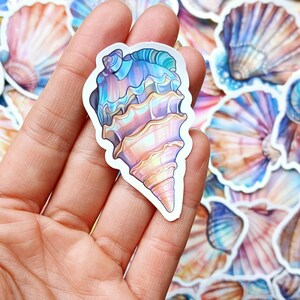 Peut inclure: Un autocollant en forme de coquillage aux couleurs bleu, violet et pêche. L'autocollant est tenu dans une main, avec d'autres autocollants de coquillages en arrière-plan. Le design est détaillé et artistique.