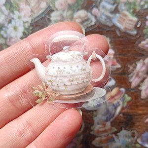 May include: A clear sticker depicting a detailed illustration of a vintage teapot. The teapot is white with floral patterns and sits on a small plate. The sticker has a transparent background, with other tea party-themed stickers visible.