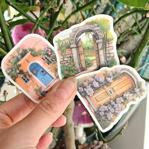 Puede incluir: Tres pegatinas troqueladas con ilustraciones de acuarela de puertas y arcos. Una pegatina muestra una puerta azul en un edificio naranja con flores rosas. Otra muestra una puerta de madera abierta en un arco de piedra. La tercera muestra una puerta de madera con flores moradas.