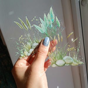 Puede incluir: Una pegatina transparente con una ilustración a la acuarela de hierbas altas, pequeñas rocas y varias flores silvestres. El diseño incorpora tonos de verde, amarillo y gris, creando un tema botánico natural. La pegatina está sujeta por una mano.