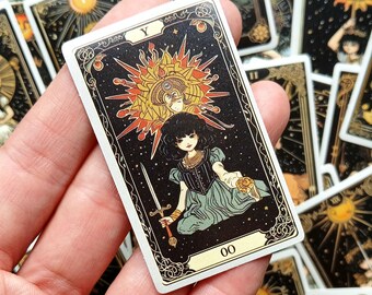 Puede incluir: Carta de tarot con fondo negro y detalles dorados. La carta representa a una mujer con cabello negro largo que lleva un vestido blanco y sostiene una espada. El número de la carta es 00 y la letra Y está en la esquina superior derecha.