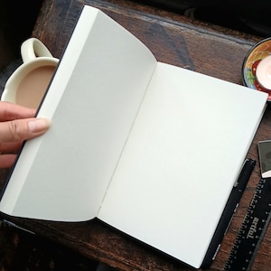 Peut inclure: Un carnet à couverture noire ouvert et vide, posé sur une surface en bois. Un stylo noir et une règle sont posés à côté du carnet.