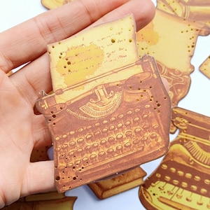 May include: A vintage-style typewriter with a gold-toned finish and a sheet of paper on top. The typewriter has a keyboard with keys and a carriage return lever. The image has a distressed, aged look with gold glitter accents.