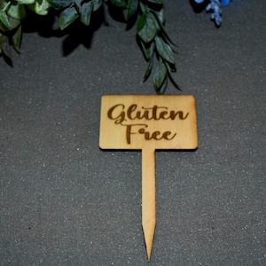 May include: A small, rectangular wooden sign with the words "Gluten Free" in a cursive font. The sign is attached to a pointed stake, suitable for placement in soil or other materials. The sign is light brown.