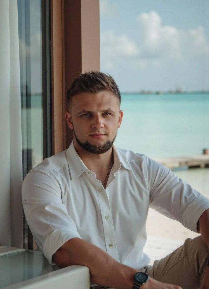 May include: A man with a beard and short brown hair is wearing a white button-down shirt and khaki pants. He is standing in front of a window with a view of the ocean.