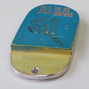May include: A metal dog tag with a blue top and a silver bottom. The blue top features a graphic of a jet and the text "SUKHOI SU-34". The tag has a small hole at the bottom.