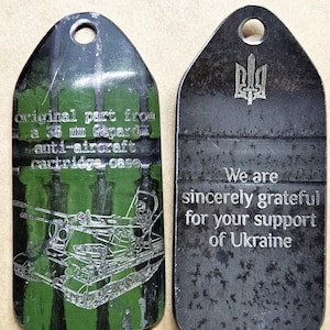 May include: Two metal tags with a hole at the top. One tag has a green and black camouflage design with text and an illustration of a tank. The other tag has the Ukrainian coat of arms and the text "We are sincerely grateful for your support of Ukraine."