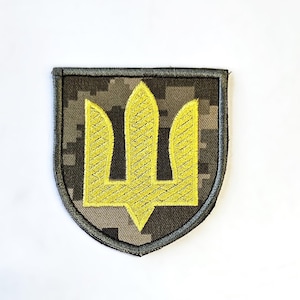 Może przedstawiać: Haftowana naszywka z ukraińskim herbem, złotym trójzębem, na tle kamuflażu. Naszywka ma kształt tarczy z szarą obwódką. Trójząb jest ważnym symbolem Ukrainy.