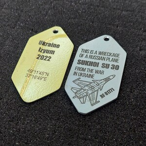 May include: Two metal dog tags. One is yellow with "Ukraine Izyum 2022" and coordinates. The other is silver with "This is a wreckage of a Russian plane Sukhoi SU 30 from the war in Ukraine" and "RF 81771".