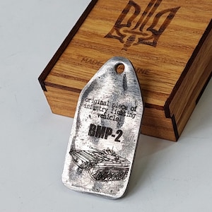 Puede incluir: Un llavero de metal plateado con una imagen en blanco y negro de un vehículo de combate de infantería BMP-2. El llavero está grabado con el texto "original piece of infantry fighting vehicle BMP-2".