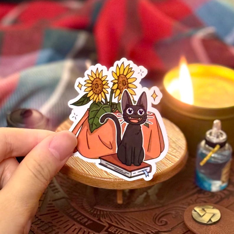 May include: A black cat with big eyes sits on a book with a sunflower bouquet in a pot behind it. The sticker is on a wooden table.