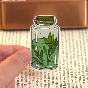May include: A sticker featuring a cartoon illustration of a glass jar with a green liquid and sprigs of green herbs inside. The jar has a cork lid and is outlined in black. The sticker is held by a hand.