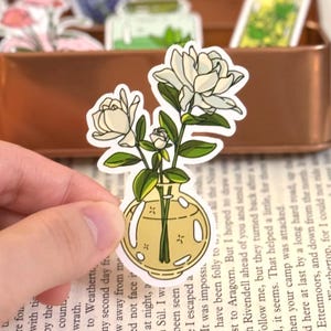 May include: A sticker featuring a floral design. The sticker depicts white flowers with green leaves in a yellow vase. The sticker is held by a hand.