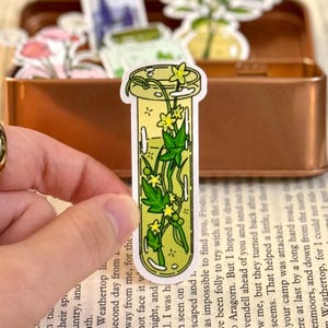 May include: A sticker featuring a yellow potion bottle with green leaves and yellow flowers inside. The sticker is held by a hand, with a book and a metal tin in the background. The sticker has a white border.