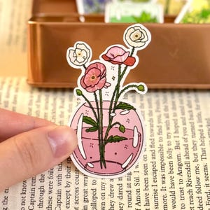 May include: A sticker featuring a pink vase with pink and white flowers. The sticker has a pink outline and is held by a hand. The background includes a book and a brown container.