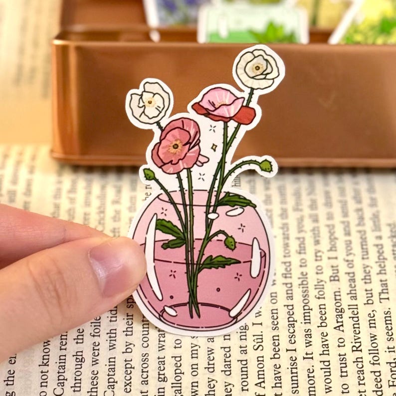 May include: A sticker featuring a pink vase with pink and white flowers. The sticker has a pink outline and is held by a hand. The background includes a book and a brown container.
