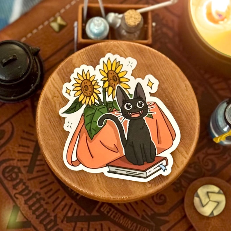 May include: A sticker featuring a black cat with bright green eyes sitting on a book next to a potted sunflower. The cat is smiling and has a surprised expression. The sticker is on a wooden surface.