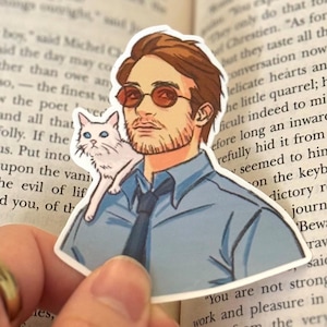 May include: A sticker featuring a cartoon illustration of a man with brown hair and a beard wearing a blue shirt, a black tie, and sunglasses. A white cat with blue eyes is perched on his shoulder. The man has a serious expression on his face.