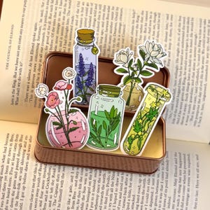 May include: A collection of decorative stickers featuring various floral arrangements in glass containers. The stickers showcase different flowers and greenery in vases and jars, with a vintage aesthetic. The stickers are arranged on a book.
