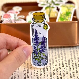 May include: A sticker featuring a clear glass bottle with a cork stopper. Inside the bottle are purple lavender flowers. The bottle has a gold charm and is held in a person's hand. Other stickers are in the background.