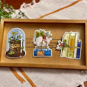 Op de afbeelding: Drie stickers op een houten dienblad. Eén sticker toont boeken onder een glazen koepel met bloemen. Een andere toont een theepot met bloemen op boeken. De laatste sticker toont boeken met een bloemig accent.