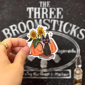 May include: A black cat with a book and sunflowers in a bright orange bag. The sticker says "The Three Broomsticks".