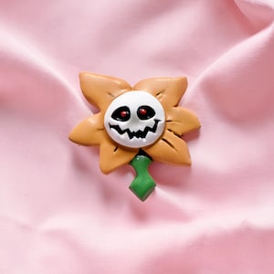 May include: A Halloween-themed flower pin with a smiling skull face at the centre. The flower has tan petals and a white face with red eyes and a jagged mouth. A green stem extends from the bottom.