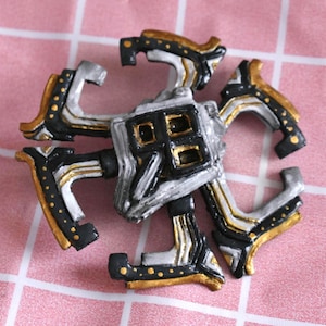 Pode incluir: Um emblema circular de várias camadas com um quadrado central e quatro recortes quadrados menores. O design é pintado em preto, prata e dourado, com quatro braços curvos que irradiam para fora, cada um acentuado com pequenos pontos dourados.
